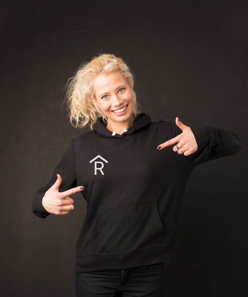 Puede incluir: Una mujer con una sudadera con capucha negra con un logotipo blanco de una casa y la letra R. Est&aacute; se&ntilde;alando el logotipo con ambas manos.