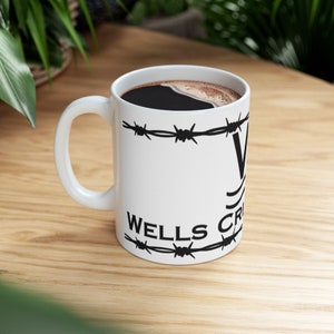 Puede incluir: Taza de cer&aacute;mica blanca con un dise&ntilde;o de alambre de p&uacute;as negro y el texto "Wells Creek" impreso en el lateral. La taza est&aacute; llena de caf&eacute;.