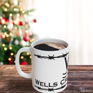 Puede incluir: Taza de caf&eacute; blanca con alambre de p&uacute;as negro y el texto "Wells Fargo" impreso en ella. La taza est&aacute; llena de caf&eacute; y est&aacute; sobre una superficie de madera.