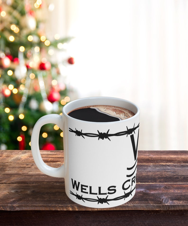 Puede incluir: Taza de caf&eacute; blanca con alambre de p&uacute;as negro y el texto "Wells Fargo" impreso en ella. La taza est&aacute; llena de caf&eacute; y est&aacute; sobre una superficie de madera.