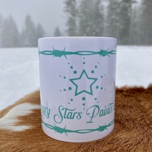 Puede incluir: Taza de cer&aacute;mica blanca con un dise&ntilde;o azul verdoso. El dise&ntilde;o presenta una estrella de cinco puntas con un c&iacute;rculo de puntos a su alrededor. El texto "Mary Stars Paints" est&aacute; escrito en una fuente cursiva debajo de la estrella. La taza est&aacute; sentada sobre una piel de vaca marr&oacute;n y blanca.