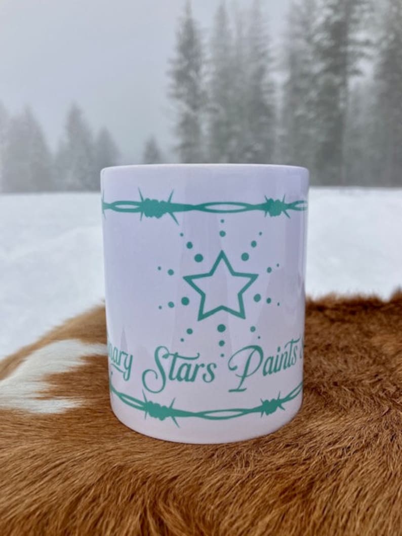 Puede incluir: Taza de cer&aacute;mica blanca con un dise&ntilde;o azul verdoso. El dise&ntilde;o presenta una estrella de cinco puntas con un c&iacute;rculo de puntos a su alrededor. El texto "Mary Stars Paints" est&aacute; escrito en una fuente cursiva debajo de la estrella. La taza est&aacute; sentada sobre una piel de vaca marr&oacute;n y blanca.