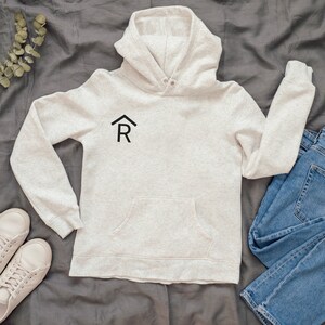 Puede incluir: Una sudadera con capucha de color gris claro con un logotipo negro de una casa y la letra R en la parte delantera. La sudadera tiene un bolsillo canguro y est&aacute; colocada sobre una superficie gris.