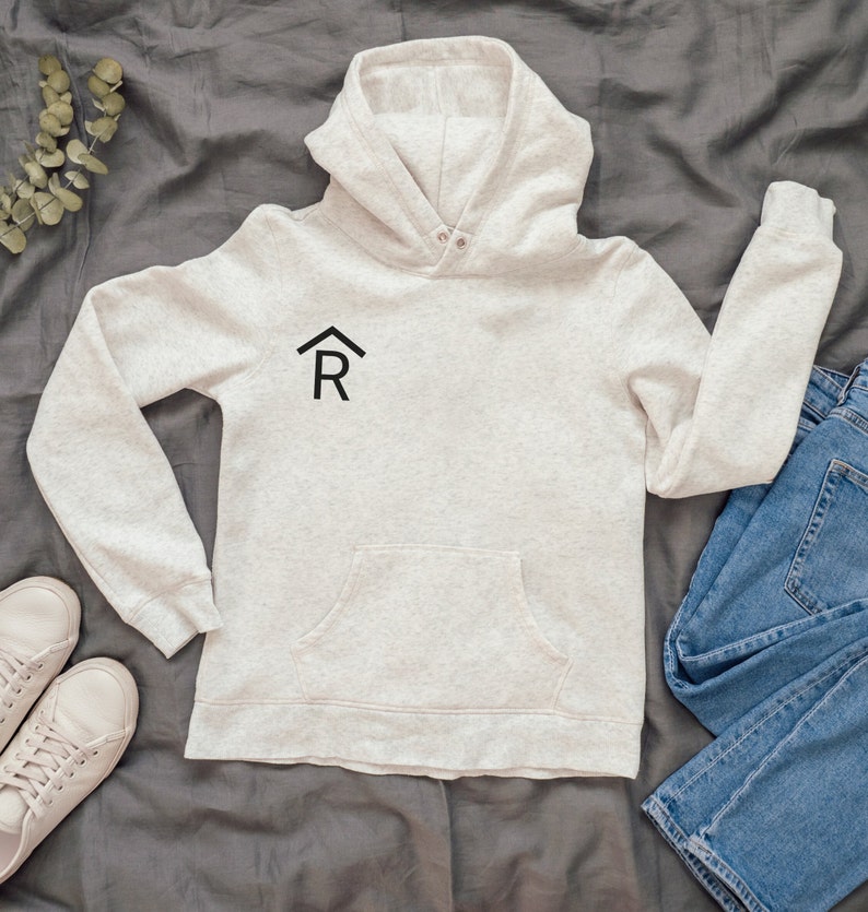 Puede incluir: Una sudadera con capucha de color gris claro con un logotipo negro de una casa y la letra R en la parte delantera. La sudadera tiene un bolsillo canguro y est&aacute; colocada sobre una superficie gris.