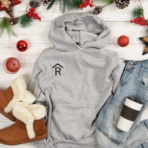 Puede incluir: Una sudadera con capucha gris con una peque&ntilde;a casa negra con la letra "R" en el interior en la parte delantera. La sudadera est&aacute; colocada sobre una superficie de madera blanca con un par de botas de piel sint&eacute;tica marr&oacute;n, unos vaqueros azules, una taza de caf&eacute; marr&oacute;n y blanca y decoraciones navide&ntilde;as.