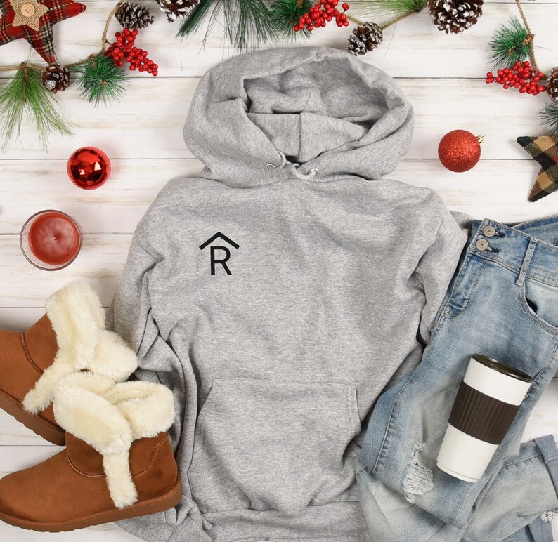 Puede incluir: Una sudadera con capucha gris con una peque&ntilde;a casa negra con la letra "R" en el interior en la parte delantera. La sudadera est&aacute; colocada sobre una superficie de madera blanca con un par de botas de piel sint&eacute;tica marr&oacute;n, unos vaqueros azules, una taza de caf&eacute; marr&oacute;n y blanca y decoraciones navide&ntilde;as.