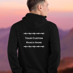 Puede incluir: Sudadera con capucha negra con un dise&ntilde;o de alambre de p&uacute;as blanco y el texto "YOUR CUSTOM RANCH NAME" en la espalda.