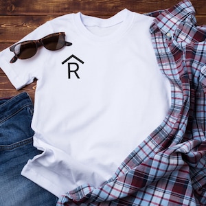 Puede incluir: Una camiseta blanca con un gráfico negro de una casa y la letra R, una camisa a cuadros azul y roja y unos vaqueros azules.