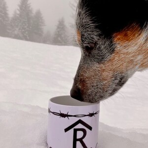 Puede incluir: Una taza de cer&aacute;mica blanca con un dise&ntilde;o de alambre de p&uacute;as negro y el texto "R Land & Co." La taza tiene una silueta negra de una casa con una "R" en su interior. Un perro blue heeler est&aacute; olfateando la taza.