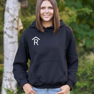 Puede incluir: Una sudadera con capucha negra con una letra "R" estilizada blanca dentro de una forma de casa en la parte delantera.