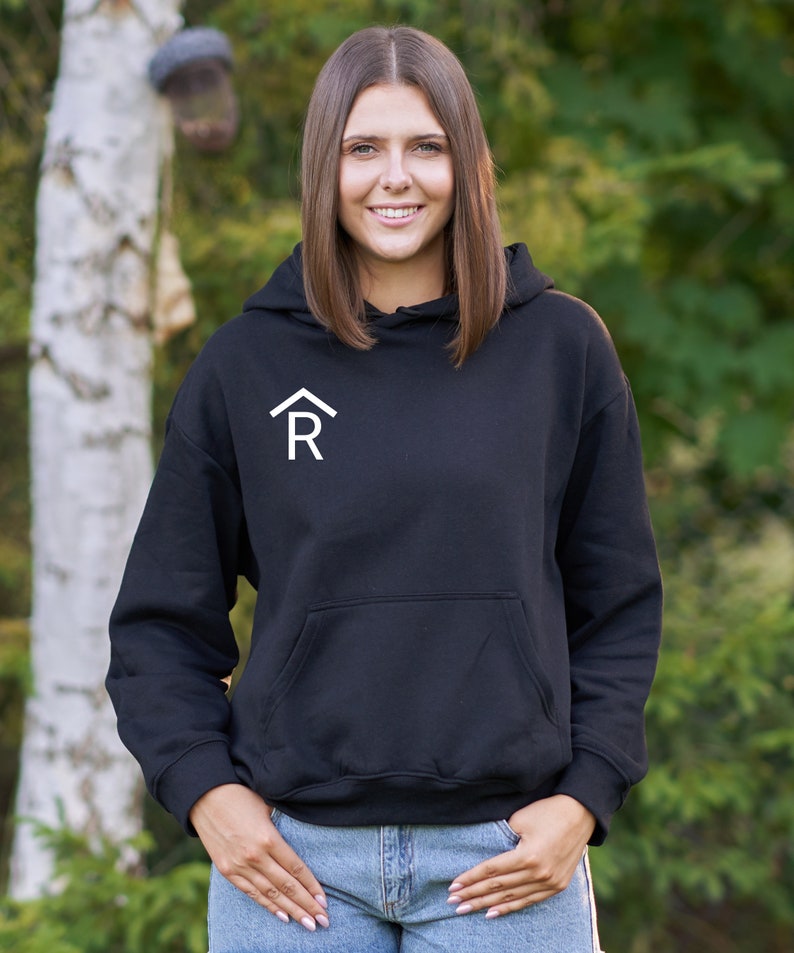 Puede incluir: Una sudadera con capucha negra con una letra "R" estilizada blanca dentro de una forma de casa en la parte delantera.