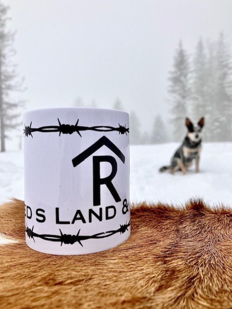 Puede incluir: Taza de cer&aacute;mica blanca con un dise&ntilde;o de alambre de p&uacute;as negro y el texto "R DS LAND &" impreso en ella. La taza est&aacute; sobre una superficie de piel marr&oacute;n.