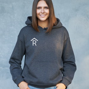 Puede incluir: Una sudadera con capucha de color gris oscuro con un logotipo blanco de una casa con una "R" en su interior. La modelo lleva la sudadera con la capucha hacia abajo y sonr&iacute;e a la c&aacute;mara.