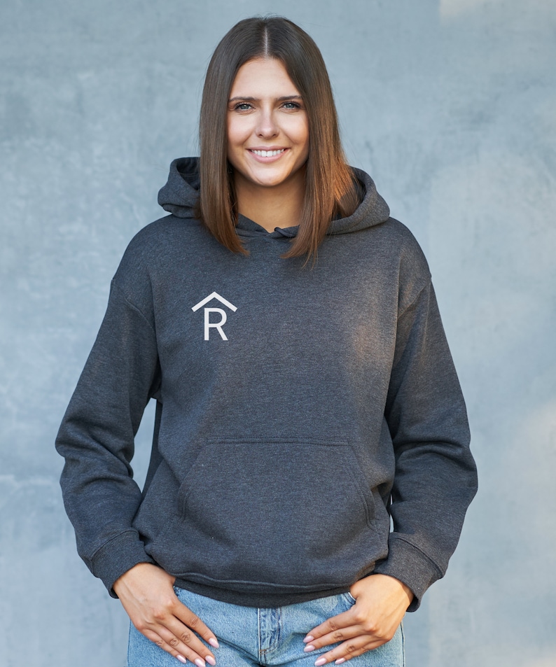 Puede incluir: Una sudadera con capucha de color gris oscuro con un logotipo blanco de una casa con una "R" en su interior. La modelo lleva la sudadera con la capucha hacia abajo y sonr&iacute;e a la c&aacute;mara.