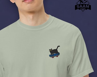 May include: A sage green t-shirt featuring a black cat skateboarding graphic. The cat has yellow eyes and is riding a blue skateboard with red wheels. The t-shirt has a crew neck and short sleeves. The text "SAAM Apparel" is printed on the t-shirt.