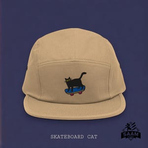 May include: A tan 5-panel cap with a black cat skateboarding graphic. The cat has yellow eyes and is riding a blue skateboard with red wheels. The text "Skateboard Cat" is printed below the graphic.