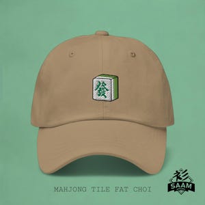 Peut inclure: Une casquette de baseball beige avec un carreau de mahjong vert brodé représentant le caractère chinois pour "gras" (發). Le carreau est entouré d'une bordure blanche. La casquette a une visière incurvée et une sangle réglable.