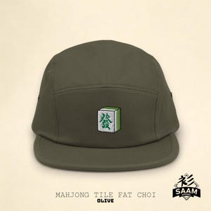 Puede incluir: Gorra de 5 paneles de color verde oliva con un azulejo de mahjong verde bordado que presenta el carácter chino para "fat choy" (prosperidad). La gorra tiene un logotipo bordado en negro que dice "Saam Apparel".
