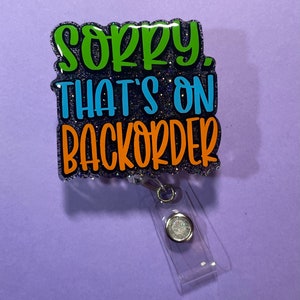 Könnte beinhalten: Ein Ausweis-Rollhalter mit dem Text "Sorry, That's on Backorder" in Grün, Blau und Orange. Das Abzeichen hat einen glitzernden schwarzen Hintergrund und einen transparenten Kunststoffclip.