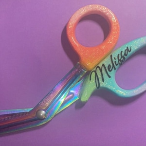 May include: Rainbow-colored medical shears with orange and blue glitter handles. The name "Melissa" is written in cursive on the blue handle. The shears are set against a purple background.