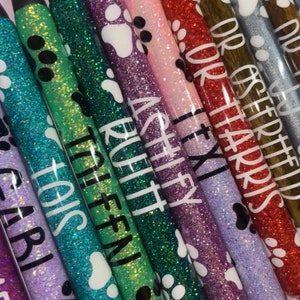 May include: A collection of glitter pens in various colors, including purple, green, and red. Each pen features a paw print design and a name in white lettering. The pens have black tips and gold-colored metal accents.