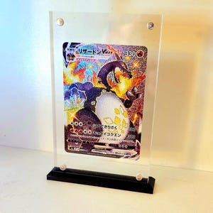 Może przedstawiać: Przezroczysta akrylowa gablota z czarną podstawą, prezentująca japońską kartę kolekcjonerską Pokemon z holograficznym obrazem postaci "リザードン VMAX" z tekstem "HP 330".