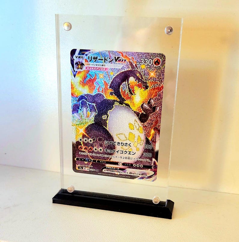 Może przedstawiać: Przezroczysta akrylowa gablota z czarną podstawą, prezentująca japońską kartę kolekcjonerską Pokemon z holograficznym obrazem postaci "リザードン VMAX" z tekstem "HP 330".