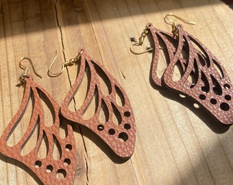 Puede incluir: Un par de pendientes de mariposa de cuero marrón con ganchos dorados. Los pendientes están recortados en forma de alas de mariposa con un diseño detallado.