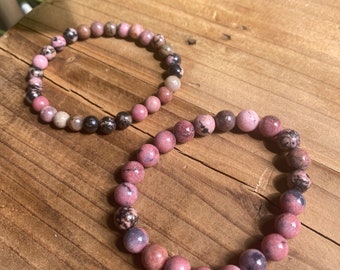 May include: Two pink and black gemstone bracelets with round beads. The bracelets are on a wooden surface.