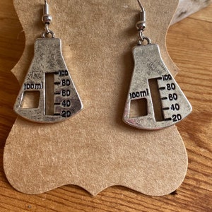 Puede incluir: Un par de pendientes de plata con forma de vaso de precipitados con una escala graduada en el lateral. Los pendientes son vasos de precipitados de 100 ml.