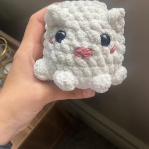 Puede incluir: Un juguete de peluche hecho a mano de ganchillo blanco en forma de cubo, con ojos de botón negros y una boca bordada en rosa. El peluche tiene orejas pequeñas y redondeadas y pies pequeños y redondeados. El juguete se sostiene en una mano.
