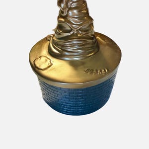 May include: A cylindrical container with a dark blue brick-patterned base and a metallic gold lid. The lid features a sculpted, weathered, pointed hat with deep folds and creases, resembling a classic wizarding hat with a distinct face-like structure.