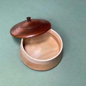 May include: A wooden box with a lid, partially open. The lid is a dark brown, and the box itself is a lighter wood. The box is round and appears to be handmade, suitable for storing small items. The box's diameter is approximately 15 cm.