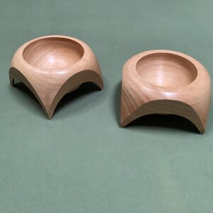 May include: Two small wooden bowls with a unique, curved, four-legged base. The bowls are made of light-coloured wood and are empty.