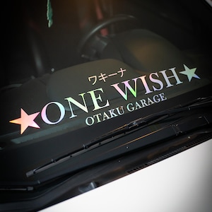 May include: A holographic vinyl decal applied to a car windshield featuring the text ONE WISH with a star on each side, the Japanese text ワキーナ above, and the text OTAKU GARAGE below. The lettering displays a multi-colored iridescent finish.