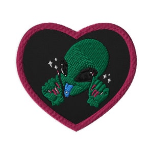 May include: Embroidered patch in the shape of a heart, featuring a green alien with black eyes and pink nails. The alien is on a black background, framed by a dark pink border. The alien has a blue tongue and is giving a peace sign.