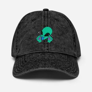 May include: A black, vintage-style baseball cap with a green alien graphic. The alien has a blue tongue, pink nails, and is winking. The cap has a curved peak and a button on top. The cap is a dark grey colour.