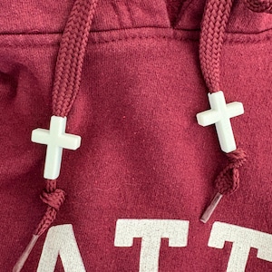 May include: Close-up of a maroon hoodie with white cross-shaped drawstring ends. The hoodie has white block lettering at the bottom. The drawstring is a thick, braided maroon cord.