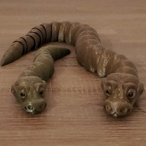 Op de afbeelding: Twee bruine, gesegmenteerde speelgoed slangen met gedetailleerde koppen en grote ogen. De slangen zijn gemaakt van een getextureerd materiaal en zijn geplaatst op een houten oppervlak.