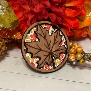 May include: A wooden maple leaf ornament with a brown wood finish. The leaf is surrounded by a circle of colorful fall leaves.