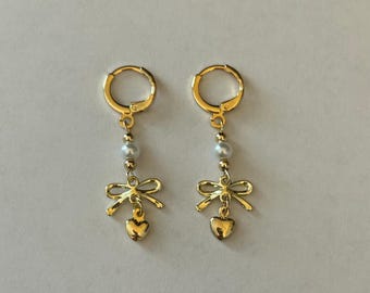 May include: A pair of gold hoop earrings with a pearl and a gold bow charm hanging from each hoop. The bow charms are attached to a small gold heart.