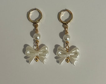 May include: A pair of gold hoop earrings with a white pearl and a white bow charm hanging from each hoop.