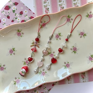 Puede incluir: Tres correas de teléfono con charms de corazón rojo y blanco, una de ellas con un charm de personaje de fresa rojo y blanco. Los charms están sobre un fondo blanco.