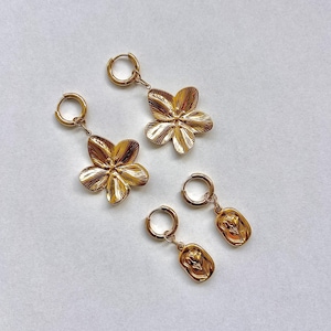May include: Gold-coloured earrings. Two pairs of earrings are displayed. One pair features a flower design, while the other has a rectangular pendant with a floral motif. Both pairs have hoop closures. The earrings are on a white background.