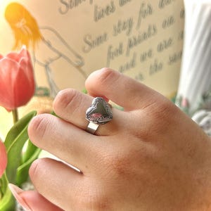 May include: A silver-toned ring with a heart-shaped design. The ring features an ornate, detailed border around the heart. The ring is on a finger, with a blurred background that includes text and a flower.
