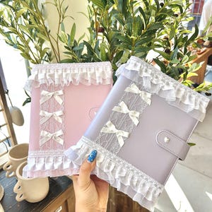 Op de afbeelding: Twee pastelkleurige agenda's met witte kanten randen en strikaccenten. Eén agenda is roze, de andere lavendel. De agenda's hebben een hartvormige sluiting en worden gepresenteerd tegen een achtergrond van groen.