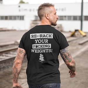 May include: A black t-shirt with the text "RE-RACK YOUR F*CKING WEIGHTS!" in white lettering. The shirt features a small graphic of a weight rack with weights on it.
