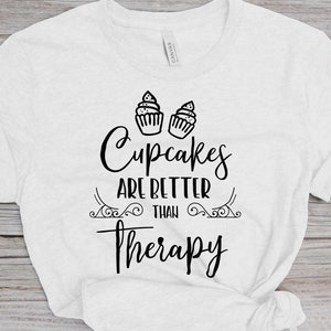 May include: White t-shirt with black text that reads "Cupcakes are better than therapy" with two cupcake illustrations above the text.