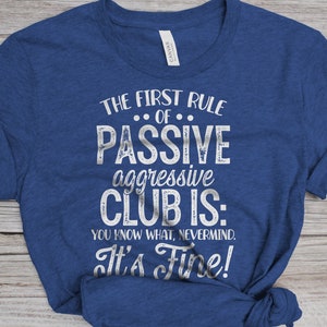 May include: A blue t-shirt with white text that reads "The First Rule of Passive Aggressive Club Is: You Know What, Nevermind. It's Fine!"