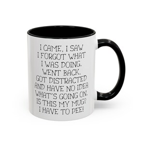 Può includere: Una tazza da caffè in ceramica bianca con bordo e manico neri. La tazza ha una scritta divertente stampata: "Sono venuto, ho visto, ho dimenticato cosa stavo facendo. Sono tornato indietro, sono stato distratto e non ho idea di cosa stia succedendo. È questa la mia tazza? Devo andare in bagno!"